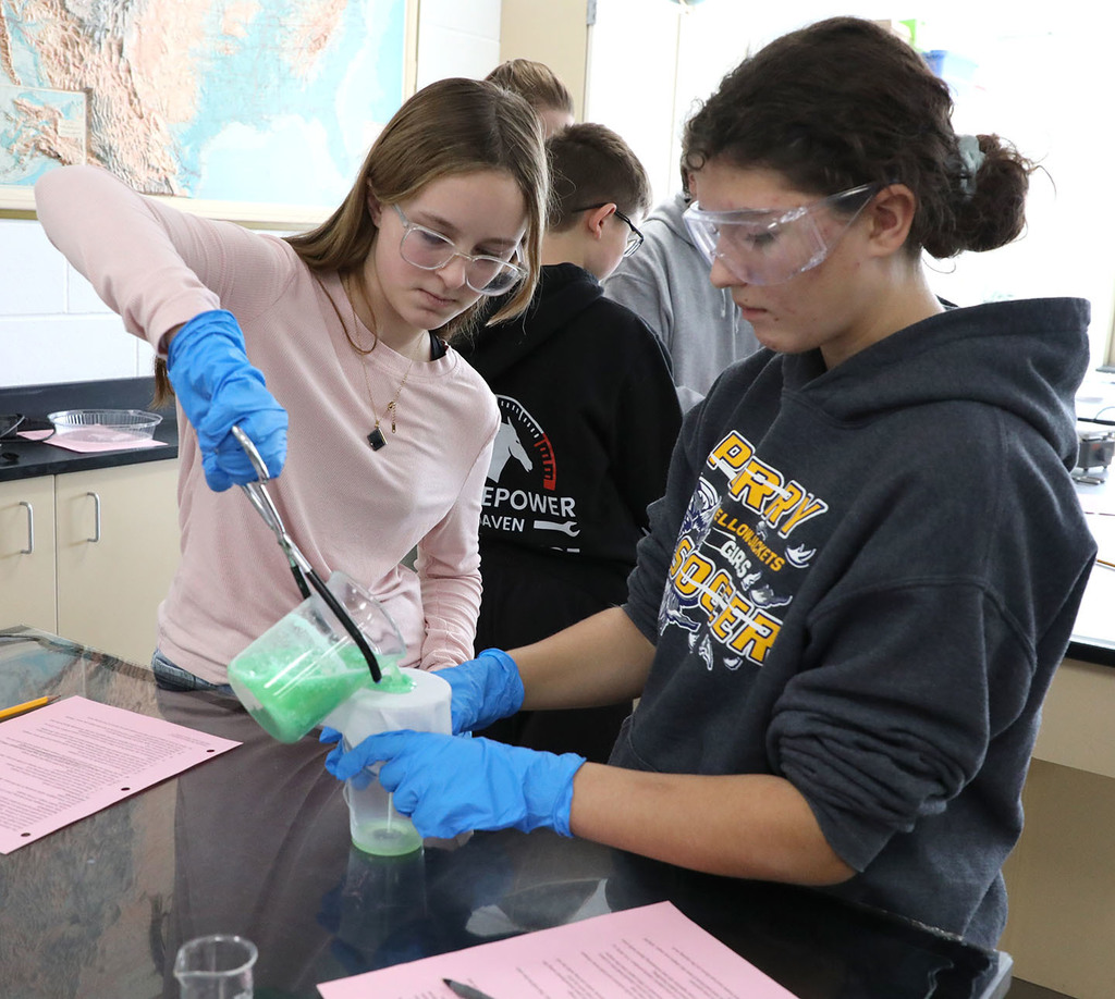 Two female Jr. High students conducting a science experiment in a classroom. You can see others in the background. 