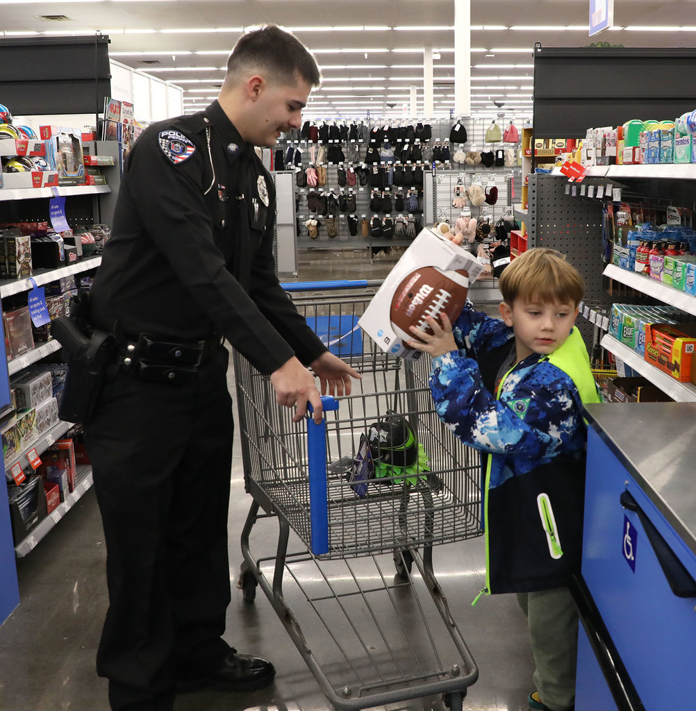 An elementary boy lifts a toy out of a cart at a store. A police officer stands next to him. 
