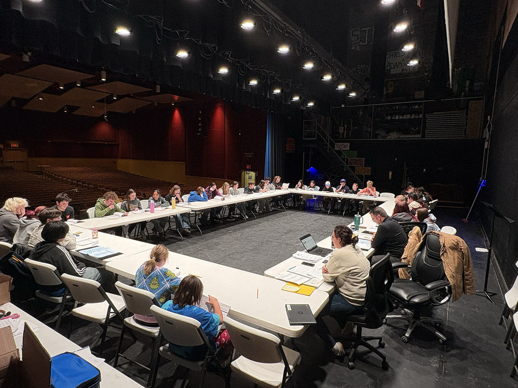 A large group of students and adults sitting around tables on an auditorium stage.