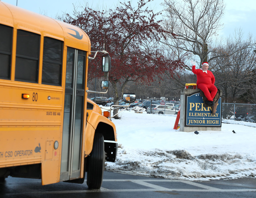 An adult male dressed as an elf. He is sitting on a school sign outside waving to a school bus. 
