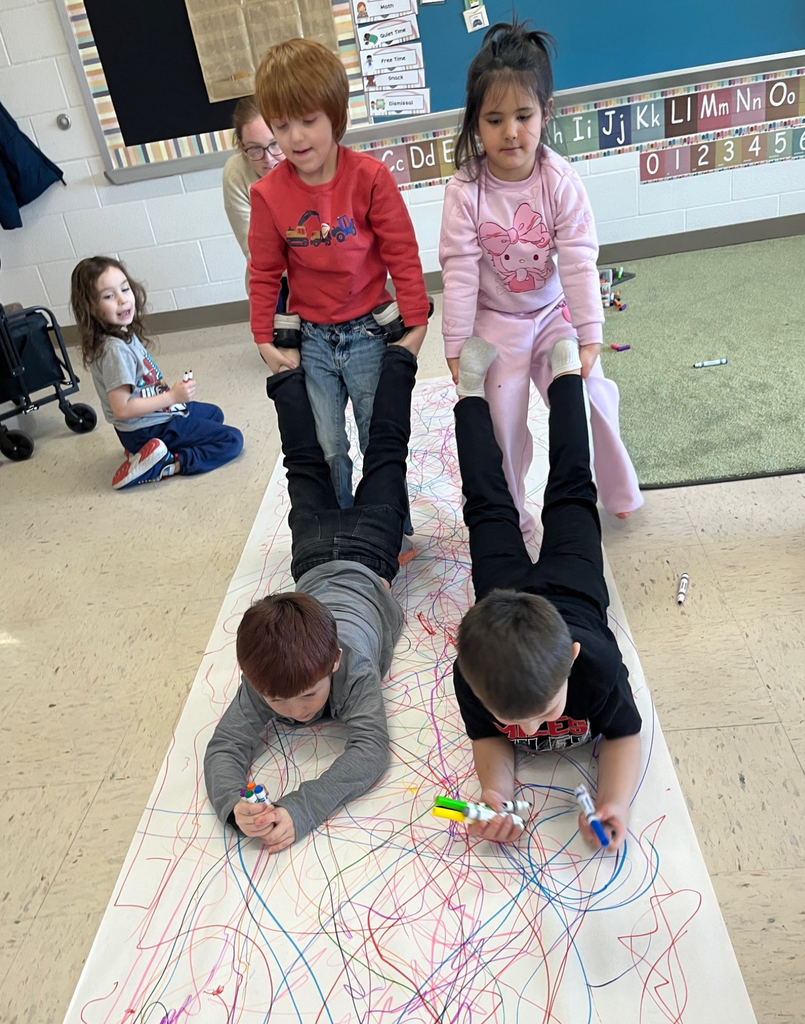 Two elementary students are holding two other students by the feet. The two students on the floor are drawing with markers. 