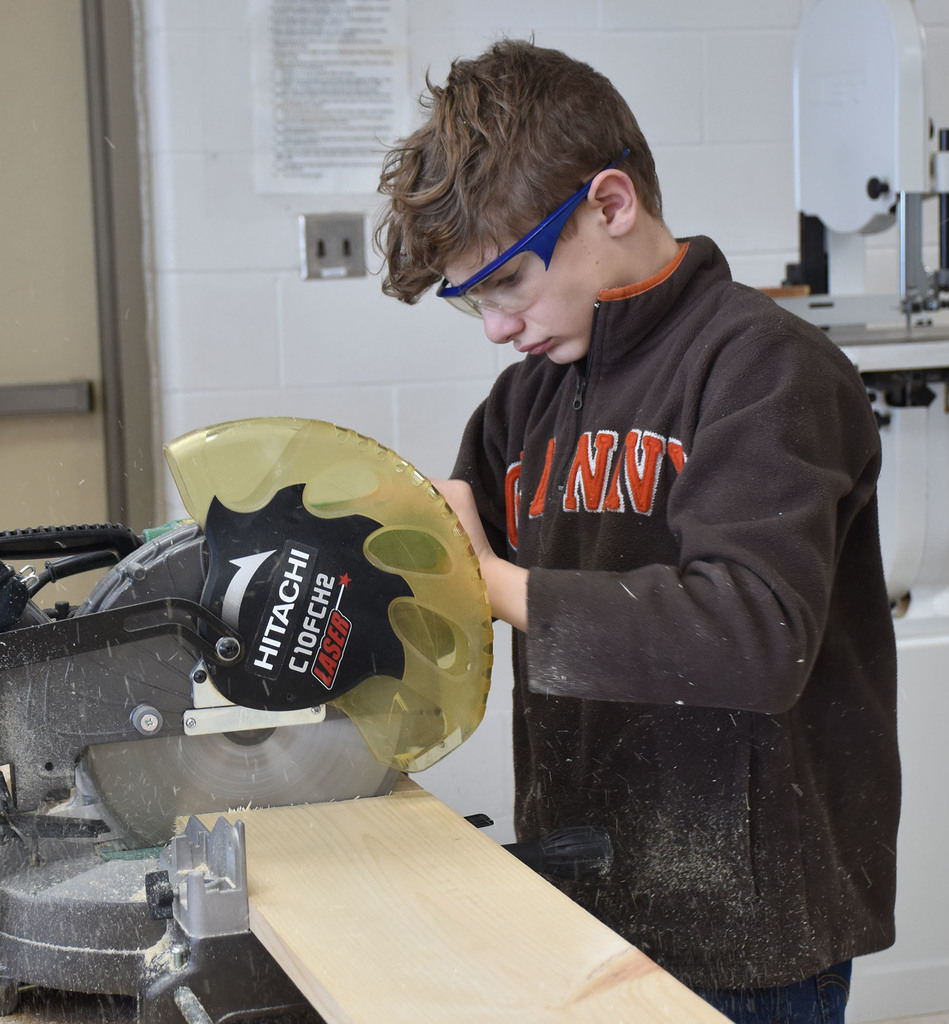 An 8th grade boy cutting a piece of wood in a school technology shop. 