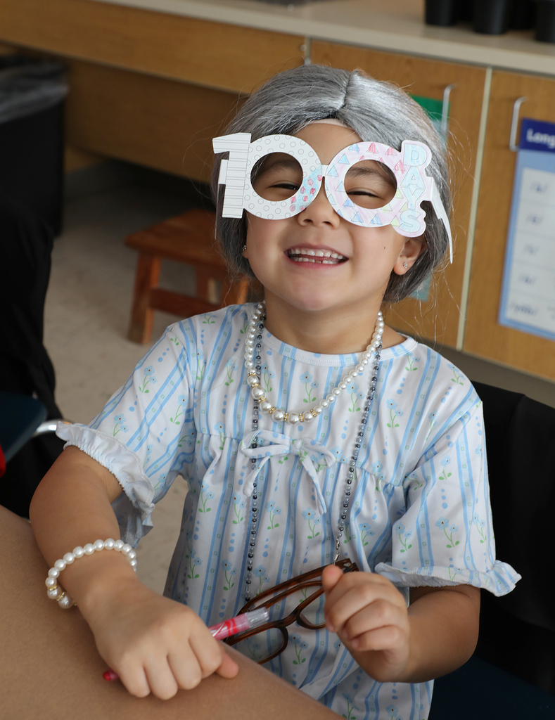A elementary girl wearing "100" glasses and a gray wig. 