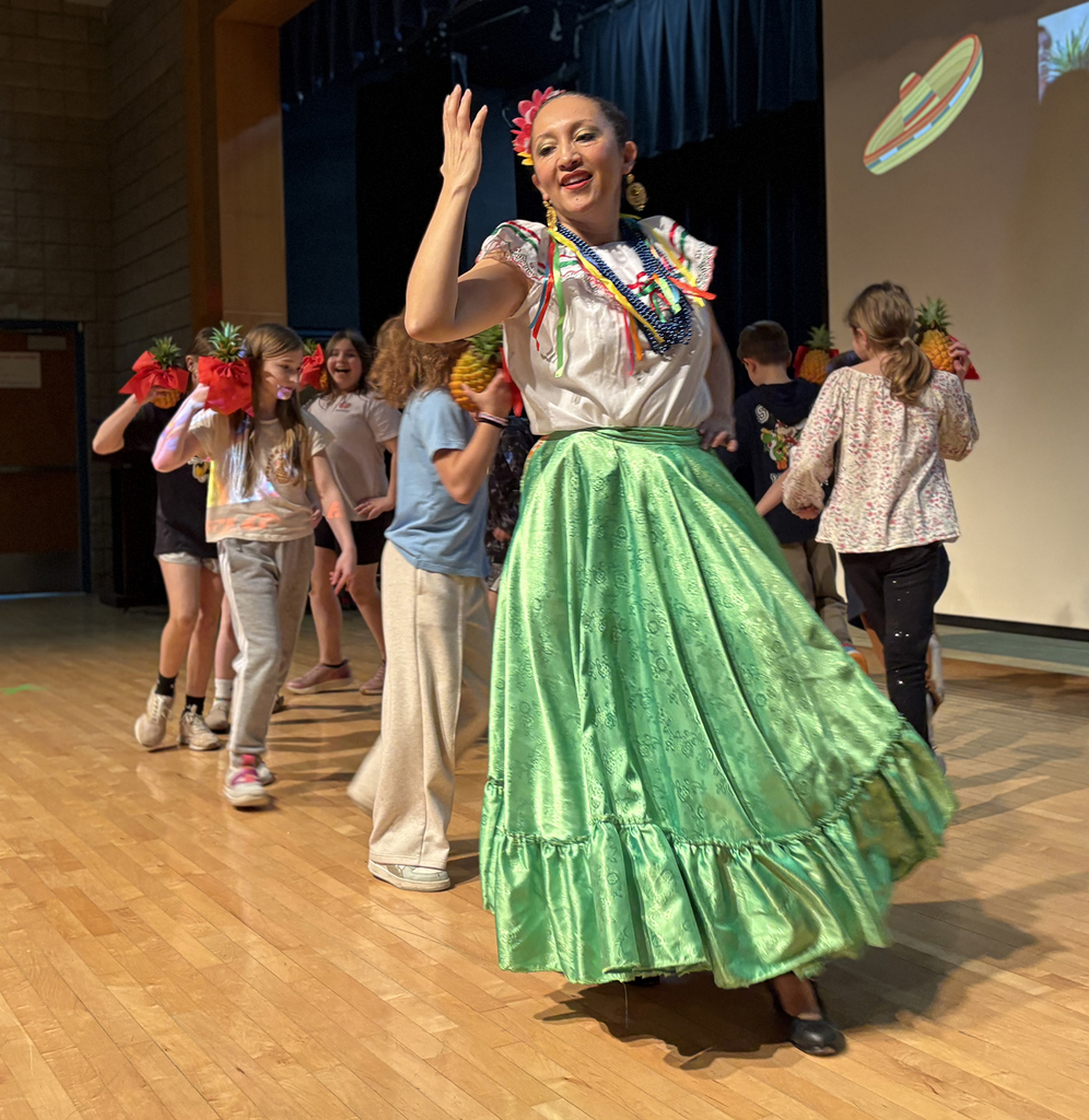 A woman in traditional Mexican attire dancing on a stage with elementary students. 