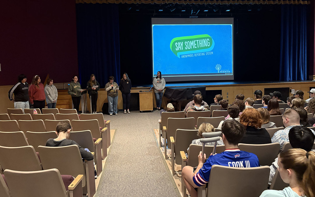 A group of high school students standing in the front of an auditorium filled with peers.