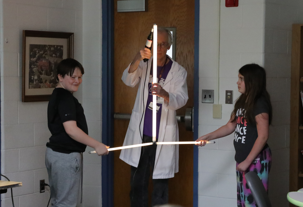 A man in a lab coat light three light bulbs. Two students are helping him. 