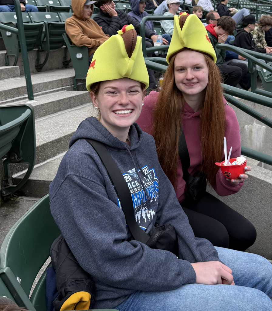 Two high school girls, with hats on, sitting in a sports stadium. 