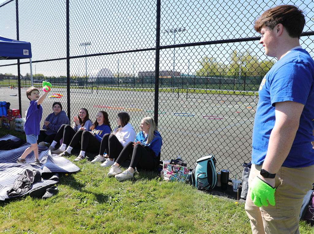 A high school student and elementary student playing catch as others look on. 