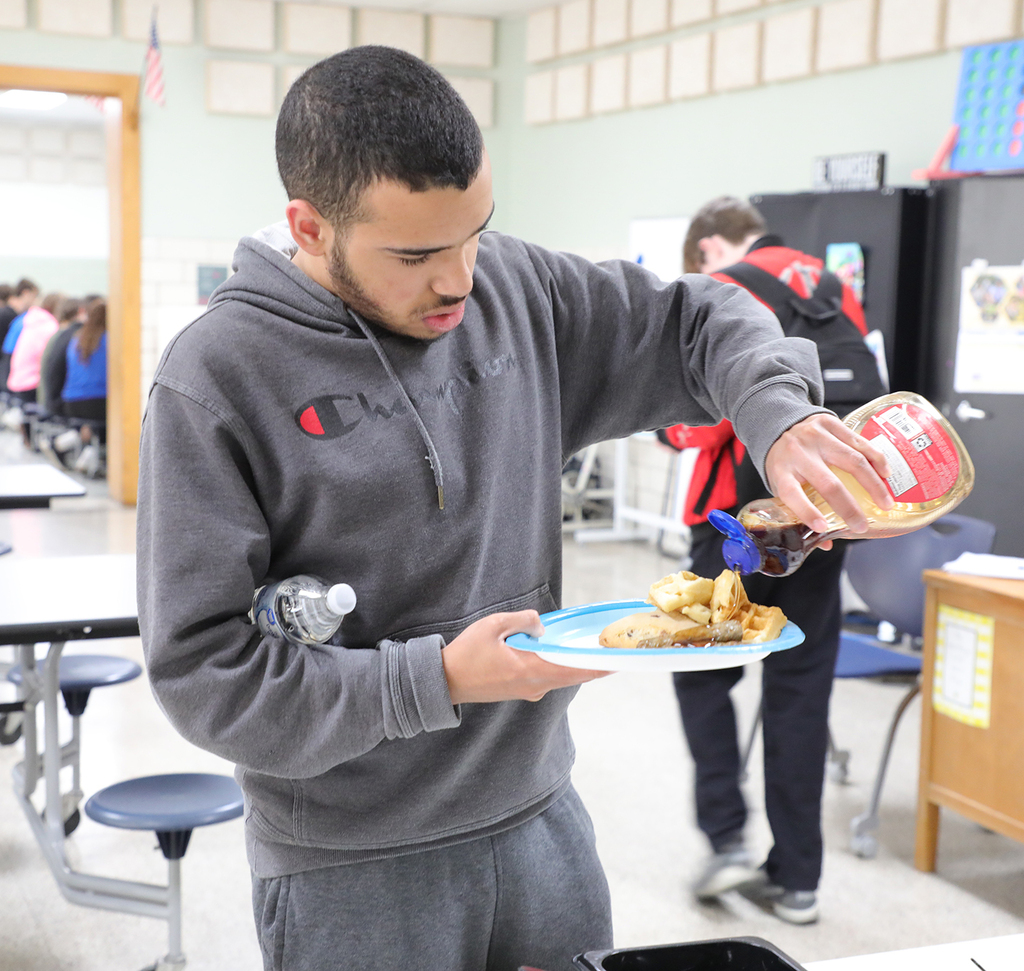 A student at a waffle station pouring syrup on his plate. 