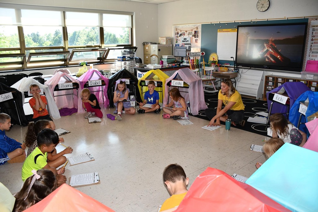 A class and teacher reading in the classroom that looks like a campsite. 
