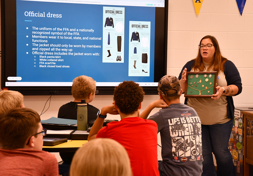 A female teacher holding up a box with medals in it. There are students in front of her. 