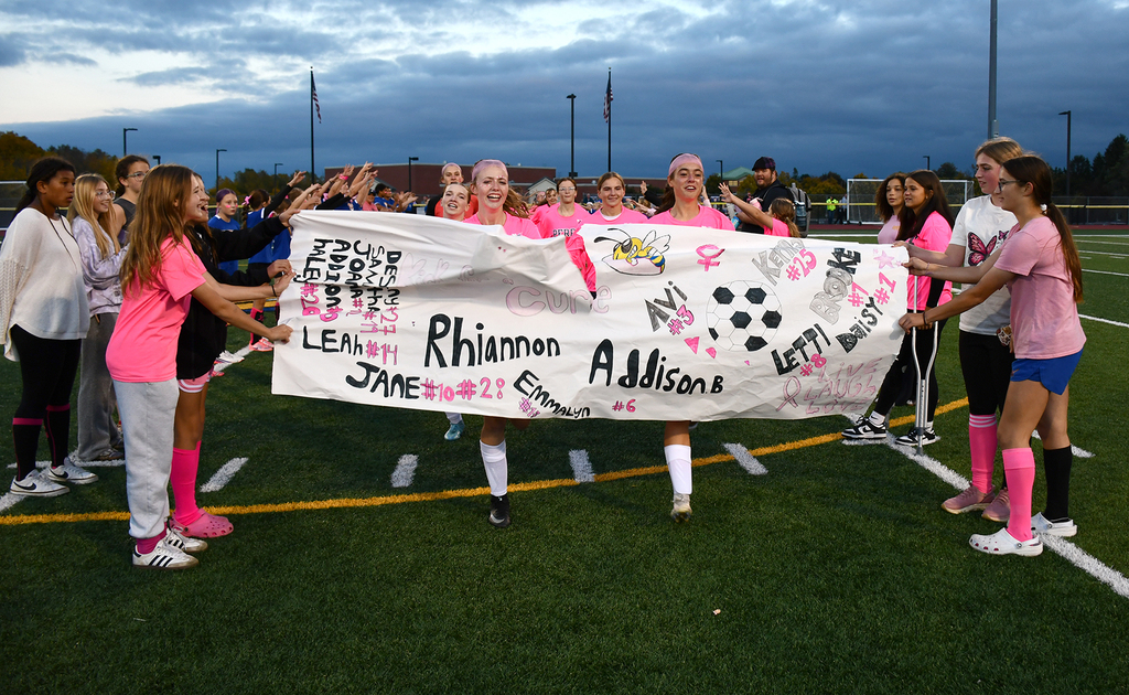 A soccer team running through a paper sign that others are holding. 
