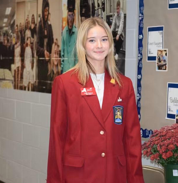 A high school girl standing in a school. She is in a red blazer.