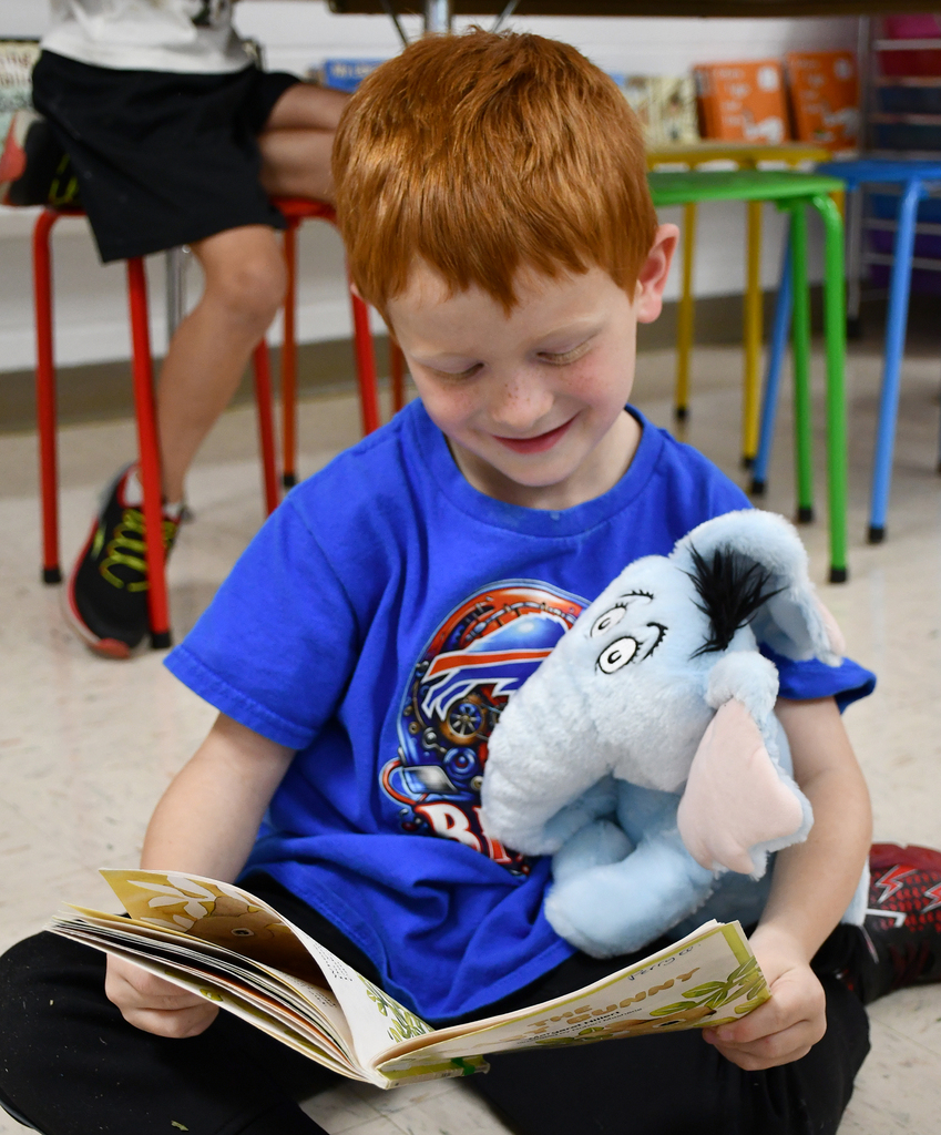 A little boy reading a book while holding onto a stuffed elephant. 