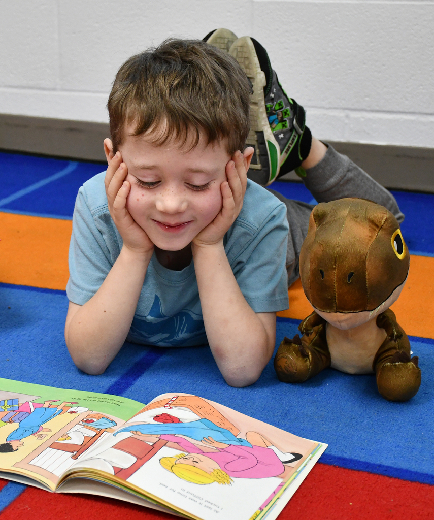 A little boy reading a book with a stuff dinosaur sitting next to him. 