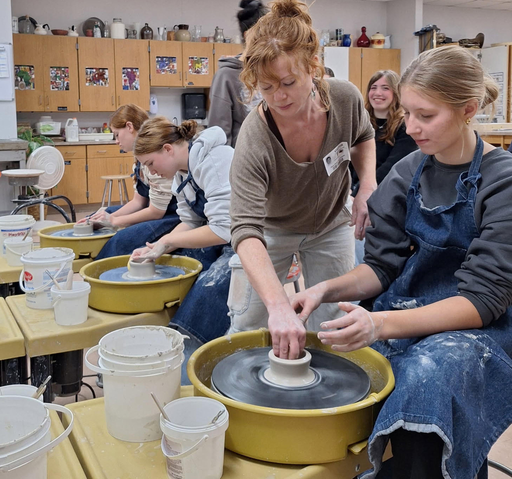 An adult female artist helping students in a high school art classroom on a pottery wheel.