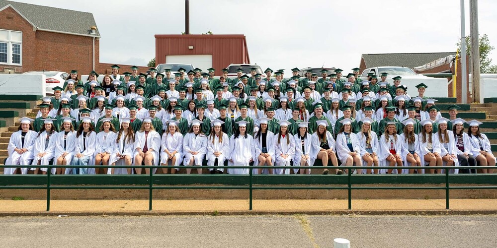 The PHS class of 2026 sits on the concrete bleachers at Pirate Stadium wearing their caps and gowns. The male graduates are in green and the female graduates in white.