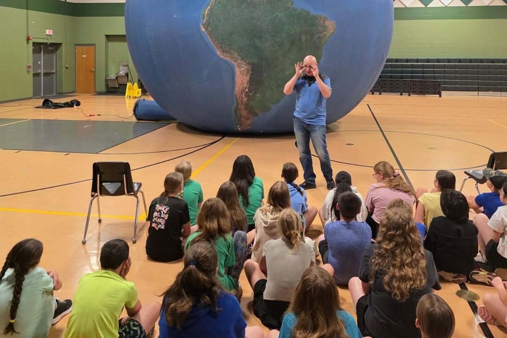 A representative from the Earth Dome planetary assembly speaks to Camp Curiosity students in the Perryville Elementary gymnasium. The planetarium is visible in the background, which looks like a large inflated globe.
