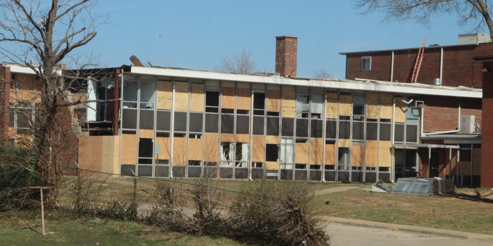 A view of the rear of the PHS building after the March 2025 tornado. There is a wall of windows that are mostly boarded up.