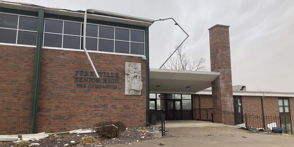 An image of the front entrance of Perryville High School shortly after the March, 2025 tornado. There is visible debris all along the ground and pieces of guttering and metal trim hanging off the roof of the building.