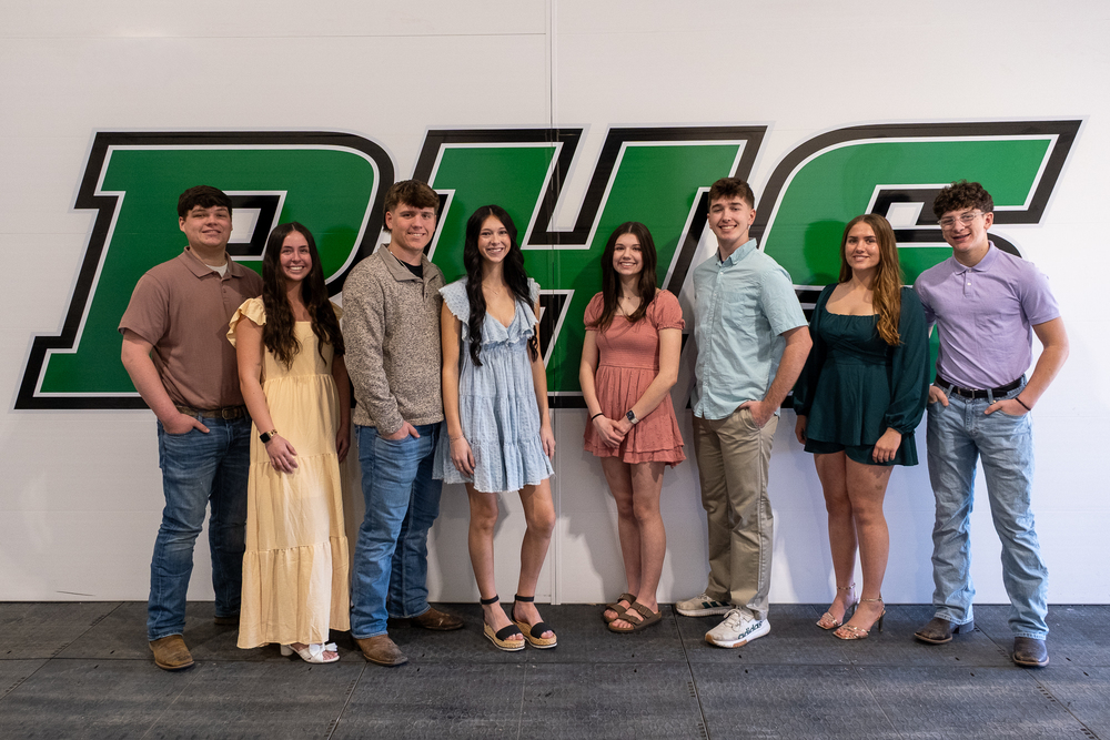 Eight members of the PHS Sweetheart Court stand in a line in front of a white wall with green PHS lettering on it.