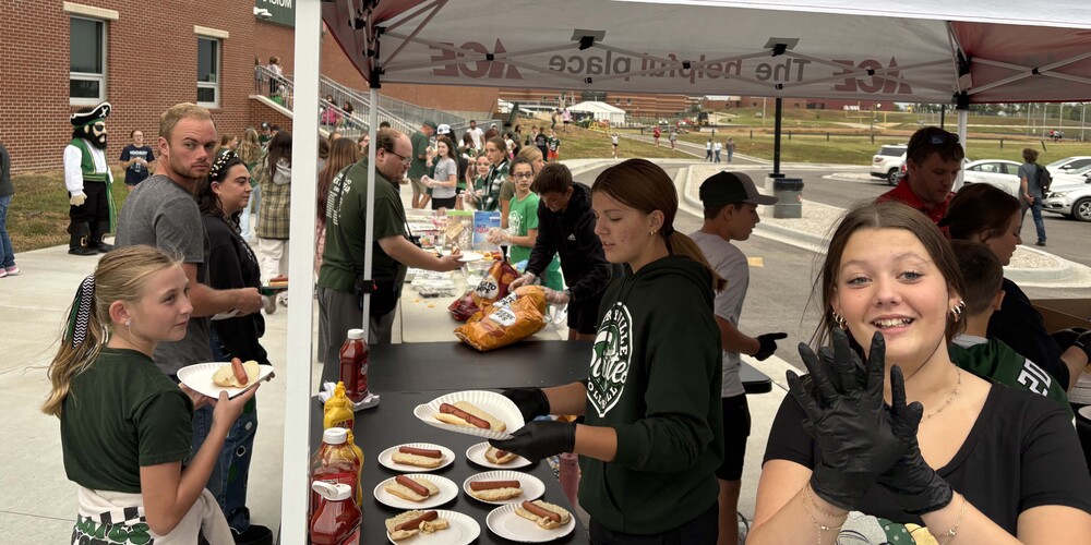 Middle school students serve hot dogs to a line of people at a tailgate picnic.