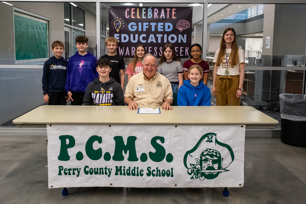 Perryville Mayor Larry Riney sits at a table with a proclamation sitting on the table. He is surrounded by PCMS gifted students. The back wall has a banner that says "Celebrate Gifted Education Awareness Week."