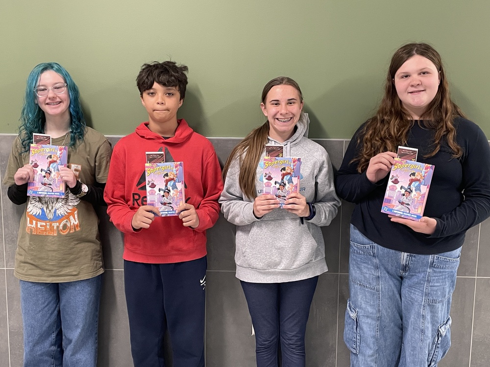 Four PCMS students stand holding a copy of the graphic novel "Supergirl"