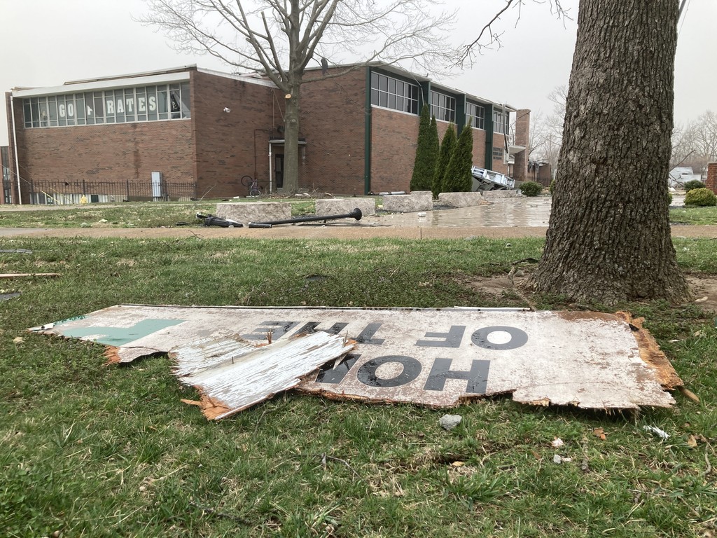 A broken sign that says "Home of the Pirates" lies in the foreground. The tornado damaged Perryville High School building is visible in the background.