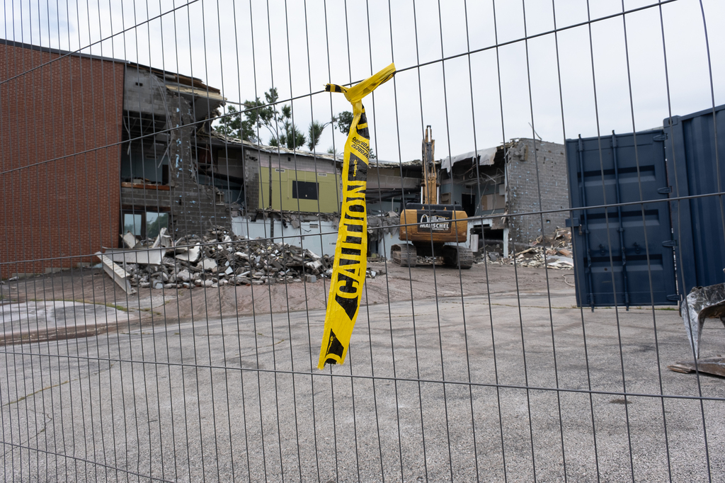 An image of yellow caution tape hanging from a fence. In the background is a backhoe doing work to demolish the damaged PHS building.