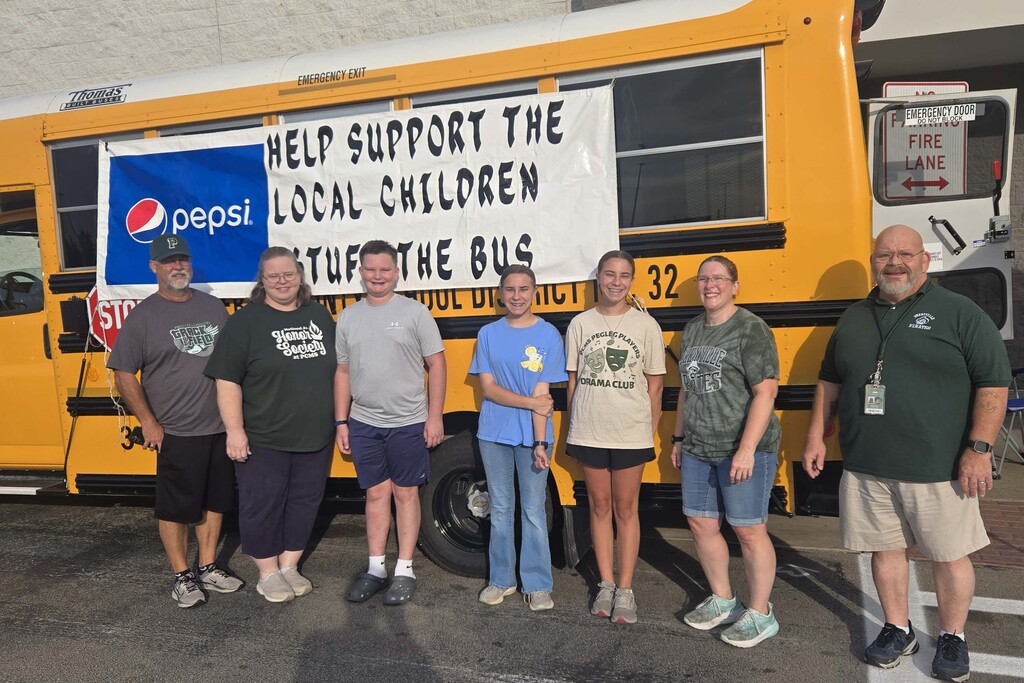 Seven people stand in front of a yellow school bus. The bus has a sign that says "Help Support the Local Children. Stuff the Bus." 