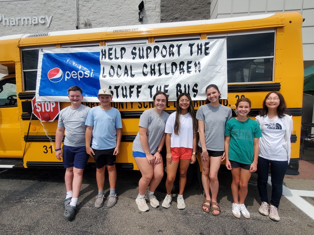 Seven students stand in front of a yellow school bus. The bus has a sign that says "Help Support the Local Children. Stuff the Bus." 