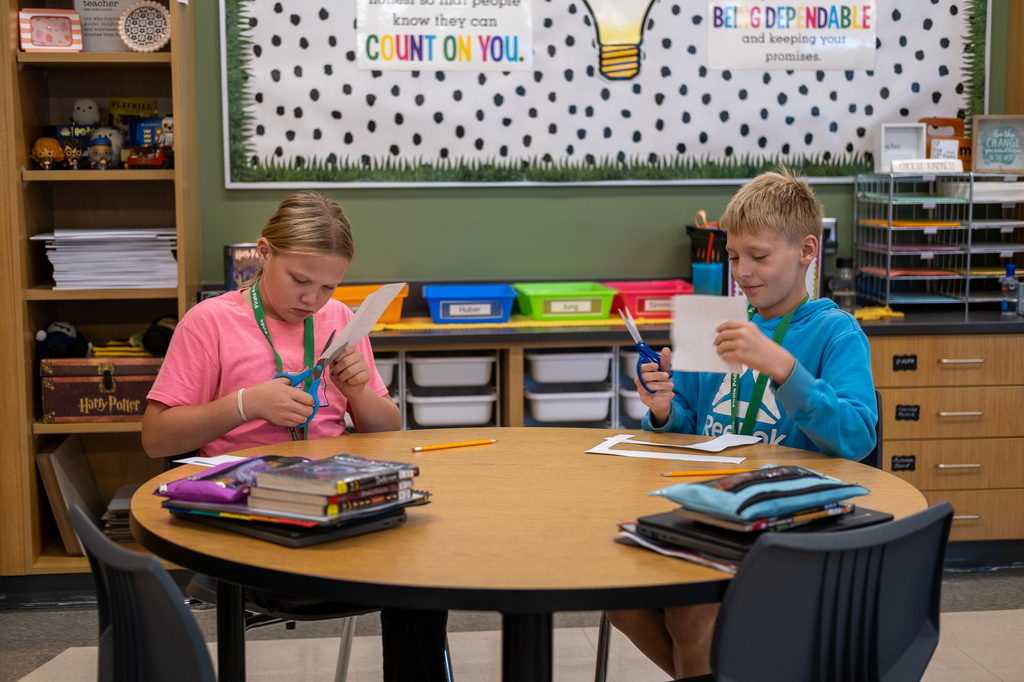 Two students sit at a desk cutting paper.