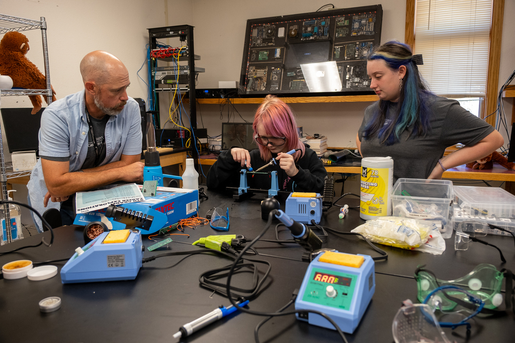 A student works on a soldering project as the instructor and another student watch.