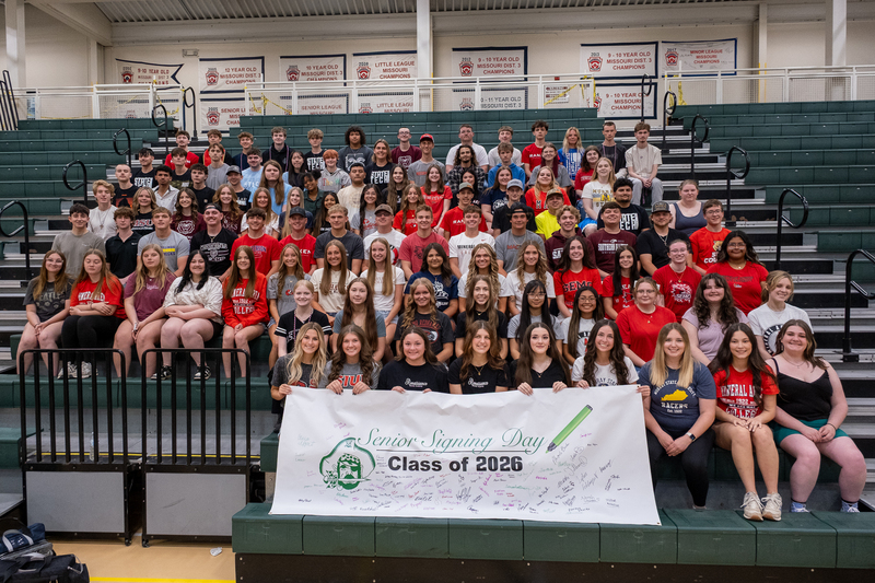 An image of all of the signing day students sitting in the bleachers at the Perry Park Center. At the front there is a banner with everyone's signatures.