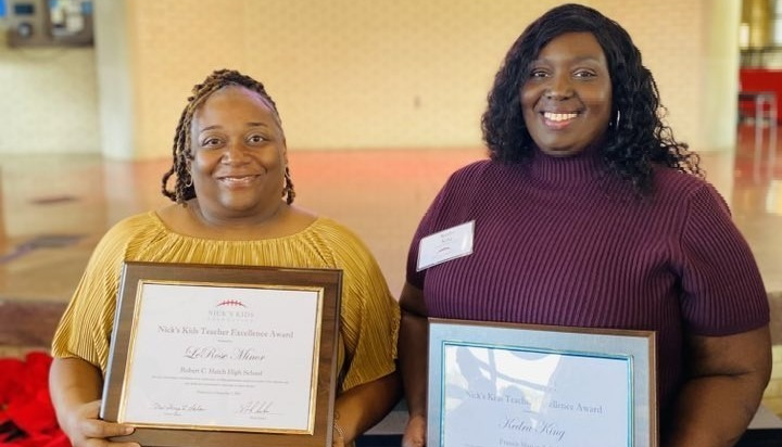 Two women stand indoors, smiling and holding framed Nick’s Kids Teacher Excellence Award certificates. The woman on the left wears a mustard yellow dress, and the woman on the right wears a maroon top. They are posed in front of a softly lit background with a poinsettia plant visible in the lower left corner.   