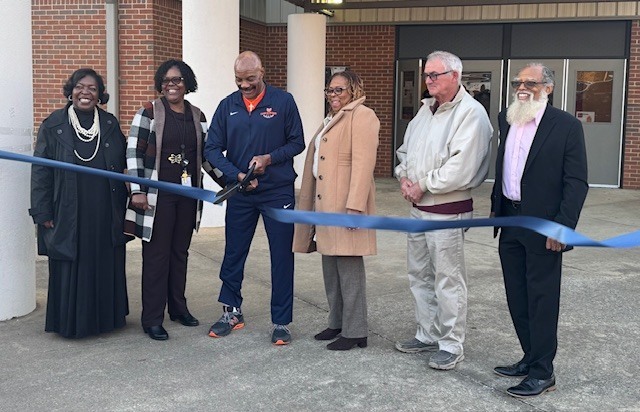 Six adults stand outside a school building holding a blue ribbon during a ribbon-cutting ceremony. One person in the center cuts the ribbon while others smile and look on. The group is dressed in professional attire, marking a celebratory school event.