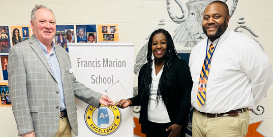 Three adults stand indoors in a school hallway next to a vertical sign that reads “Francis Marion School,” features an A+ College Ready seal, and lists “2024–2025.” The person on the left wears a blazer and holds a certificate or document, the person in the center wears a black suit and smiles, and the person on the right wears a white shirt with a striped tie. A school mural and photo display are visible in the background.