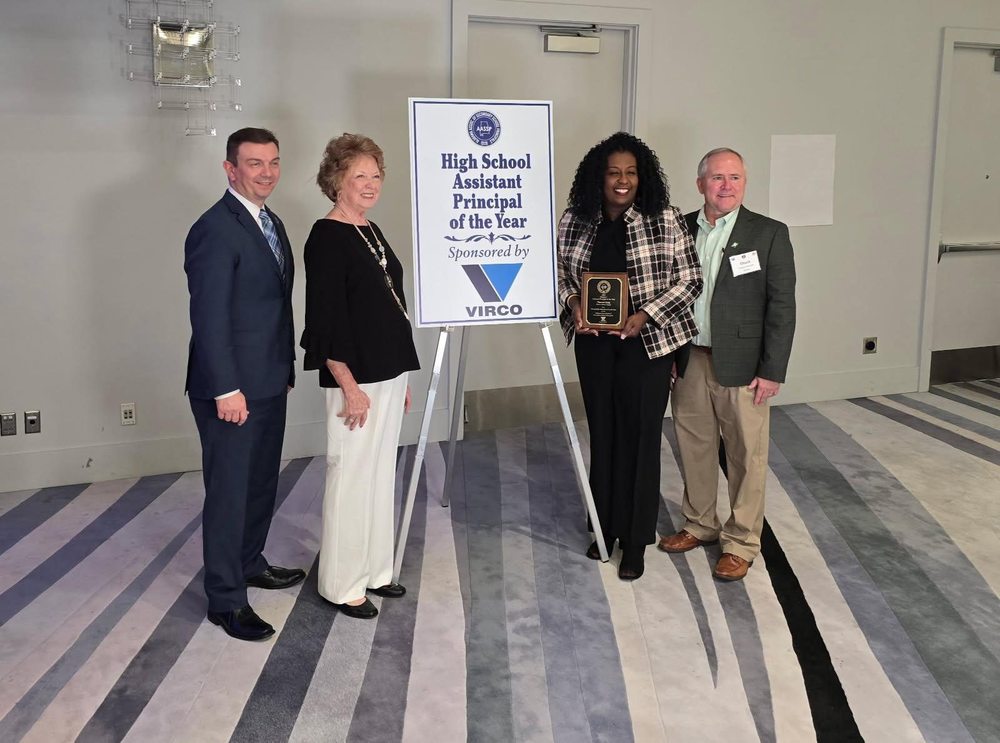 Four adults stand in a row indoors next to a large easel sign that reads “High School Assistant Principal of the Year – Sponsored by VIRCO.” The woman second from the right is holding a small rectangular award plaque and smiling. They are dressed in business attire and posed on a striped carpet in front of a plain light-colored wall.