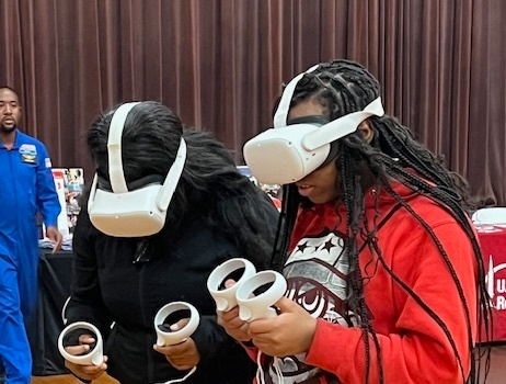 Two students wearing virtual reality headsets and holding VR controllers while participating in an interactive activity inside a gymnasium.