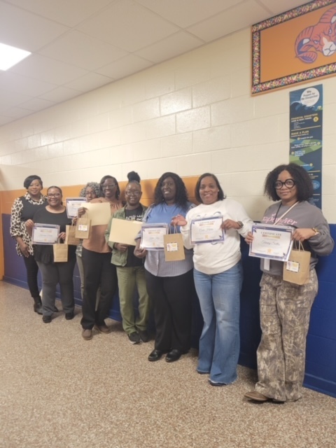 A group of eight adults standing in a school hallway, smiling and holding certificates and gift bags. They are lined up against a wall decorated with school colors and a large tiger-themed poster. The atmosphere appears celebratory, as if they have received awards or recognition.