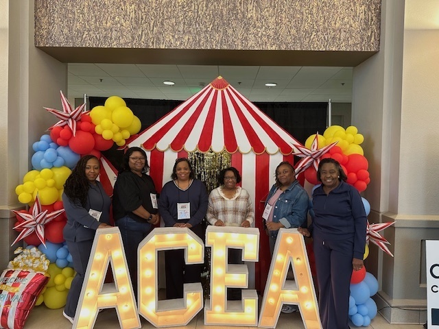 A group of six adults posing in from of a circus tent themed background at the ACEA Conference.
