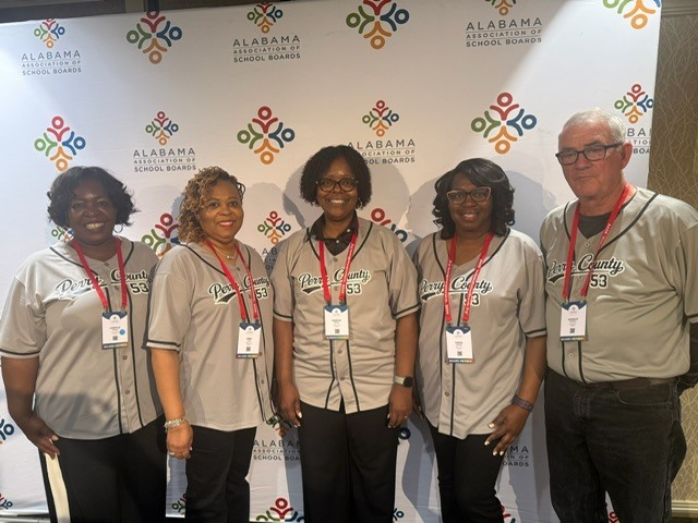 Five adults stand side by side in front of an “Alabama Association of School Boards” step‑and‑repeat backdrop, all wearing matching gray “Perry County 53” baseball-style jerseys and conference name badges, smiling toward the camera.