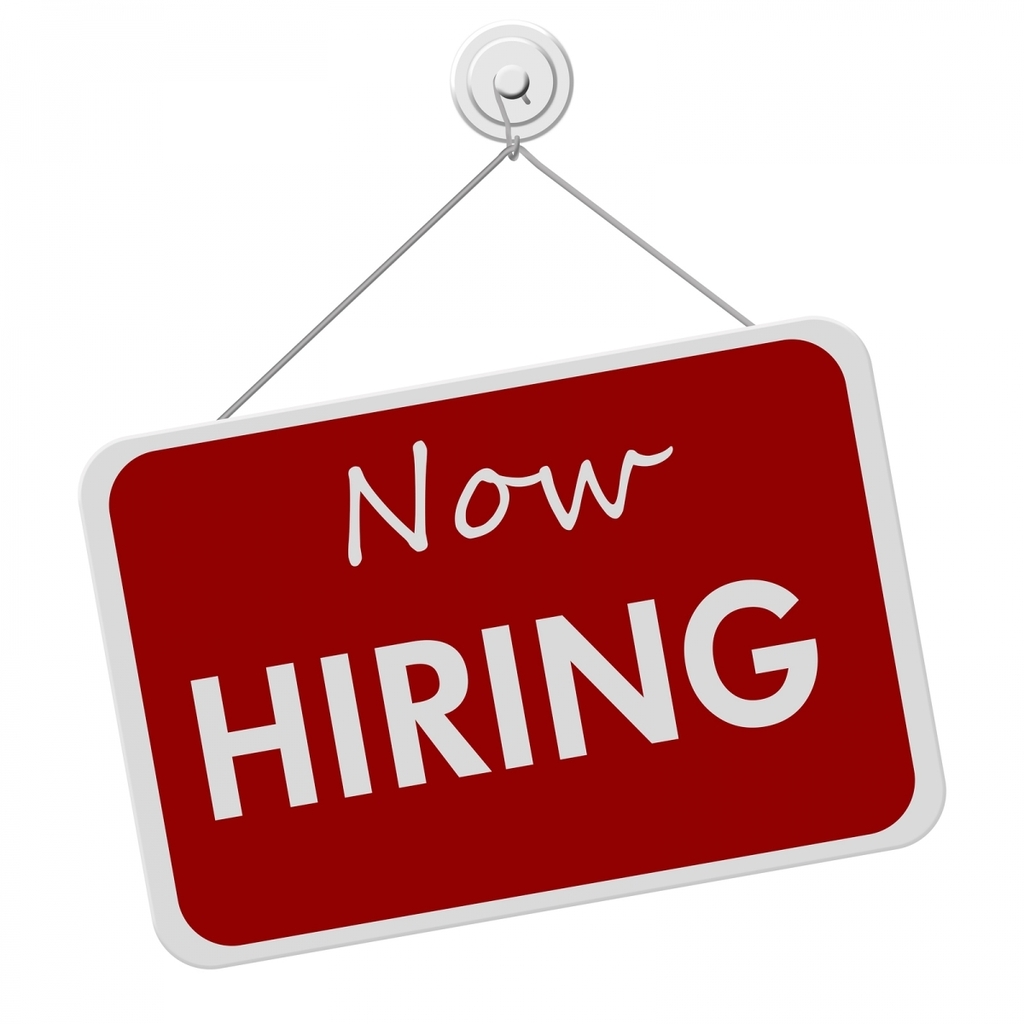 Red hanging sign with the words “Now Hiring” displayed in large white letters. The sign is suspended by a string from a wall hook and is designed to advertise employment opportunities or open job positions.
