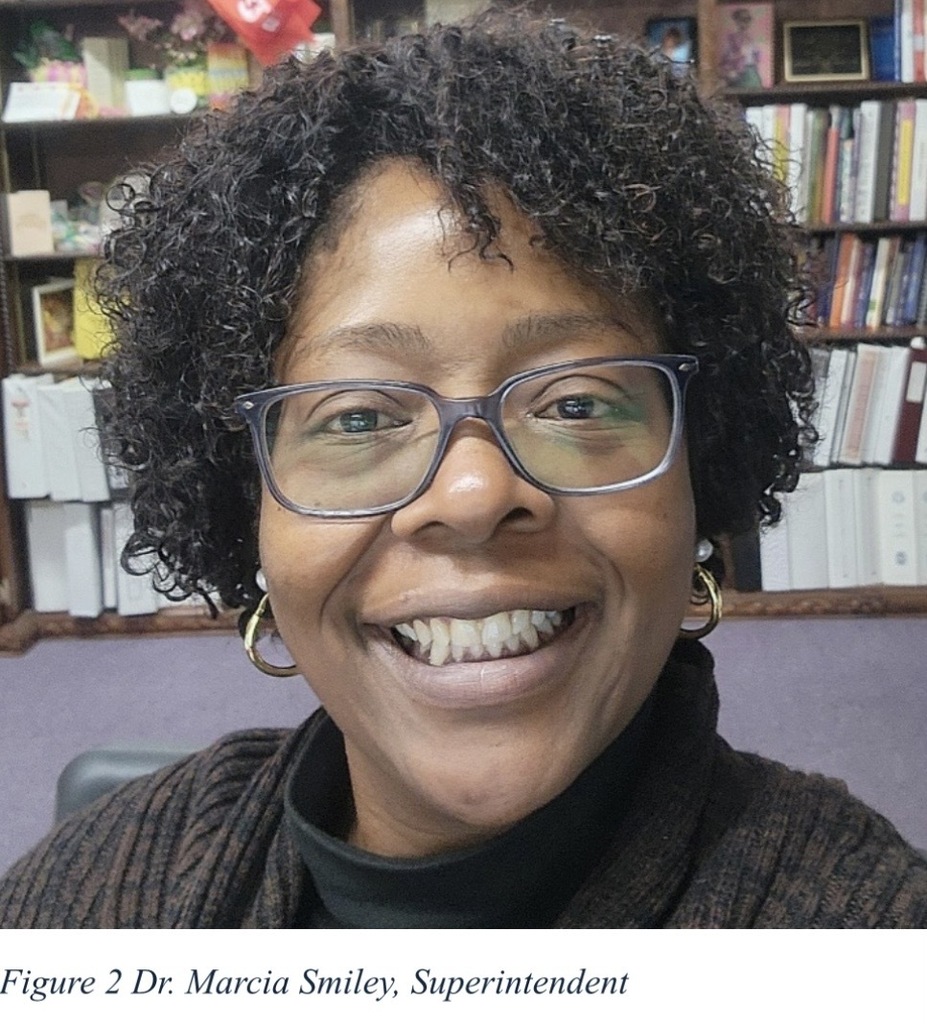 A smiling woman wearing rectangular eyeglasses is pictured in an office setting. She has short, curly hair and is wearing hoop earrings, a dark turtleneck, and a cardigan sweater. Bookshelves filled with books, binders, and office materials are visible in the background. The image appears to be a professional portrait or workplace photo. A caption below the image reads, "Figure 2 Dr. Marcia Smiley, Superintendent."