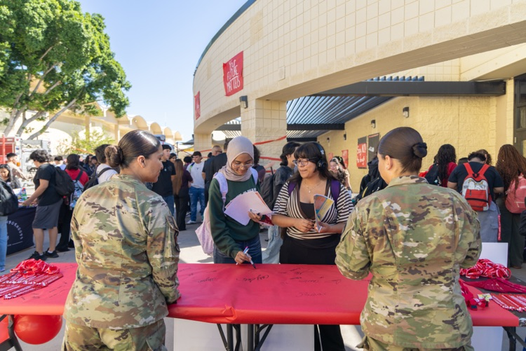 Soldiers in camouflage uniforms stand behind a red table. People stand and wait in line.