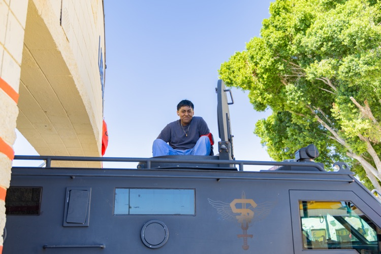 A young man wearing a gray shirt and pants sits on a black armored vehicle. Trees and a building are in the background.