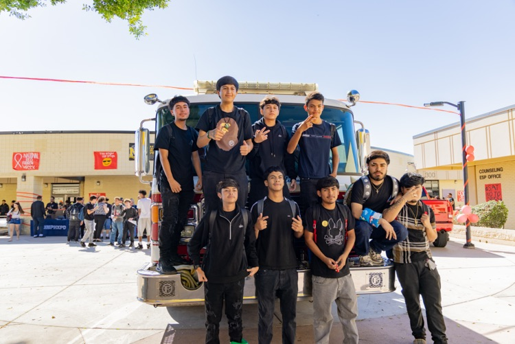 A group of students pose on the back of a fire truck with a crowd in background.