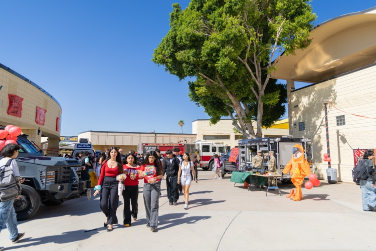 People walking on sidewalk with buildings in background. Trees, parked vehicles, and people in orange suits.