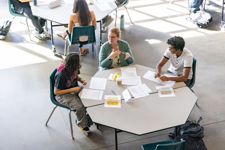 Group of four students seated around a large table, each with papers and pens, engaged in a discussion.